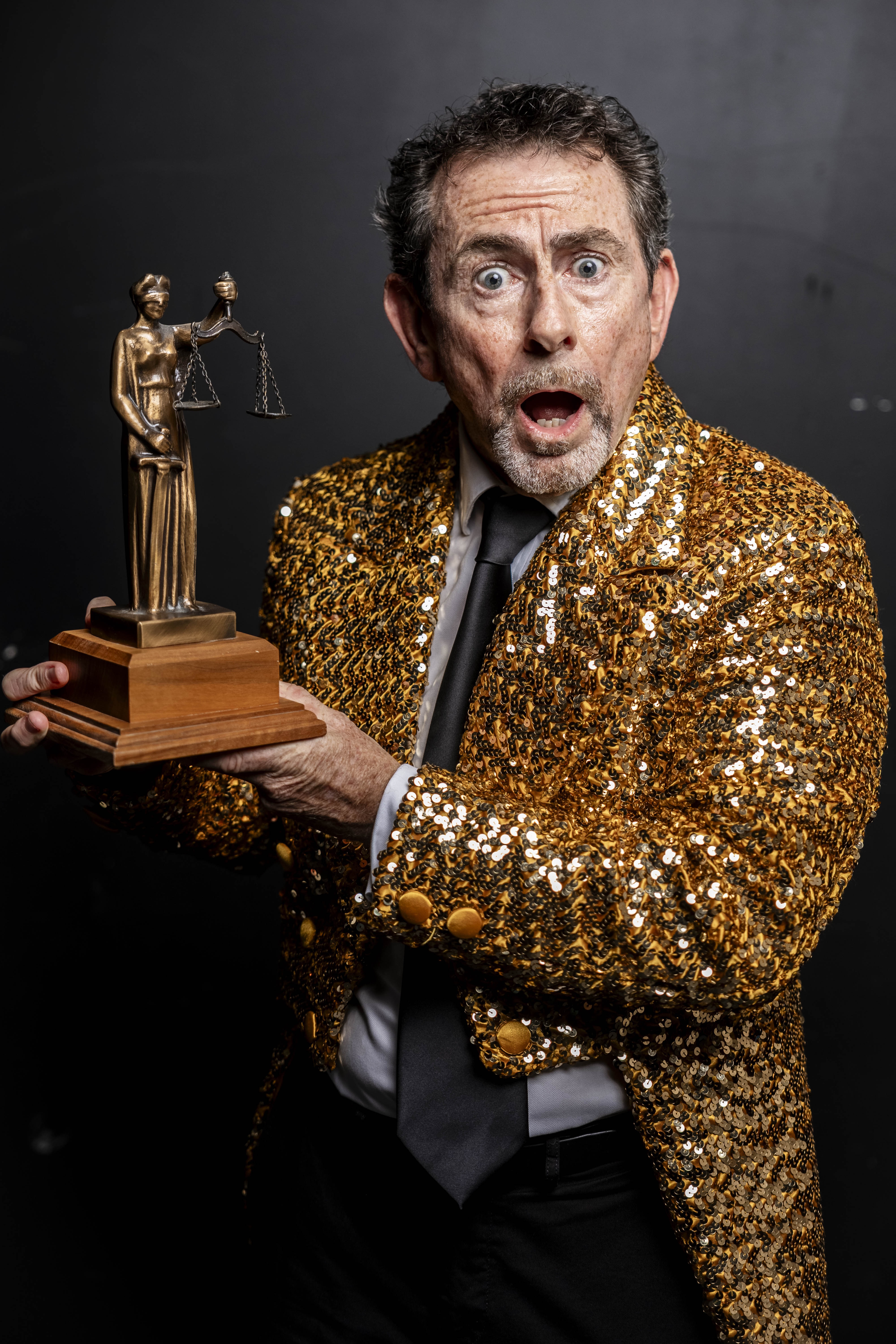 Nick Abrahams in gold sequin jacket holding a Lady Justice trophy with a surprised expression