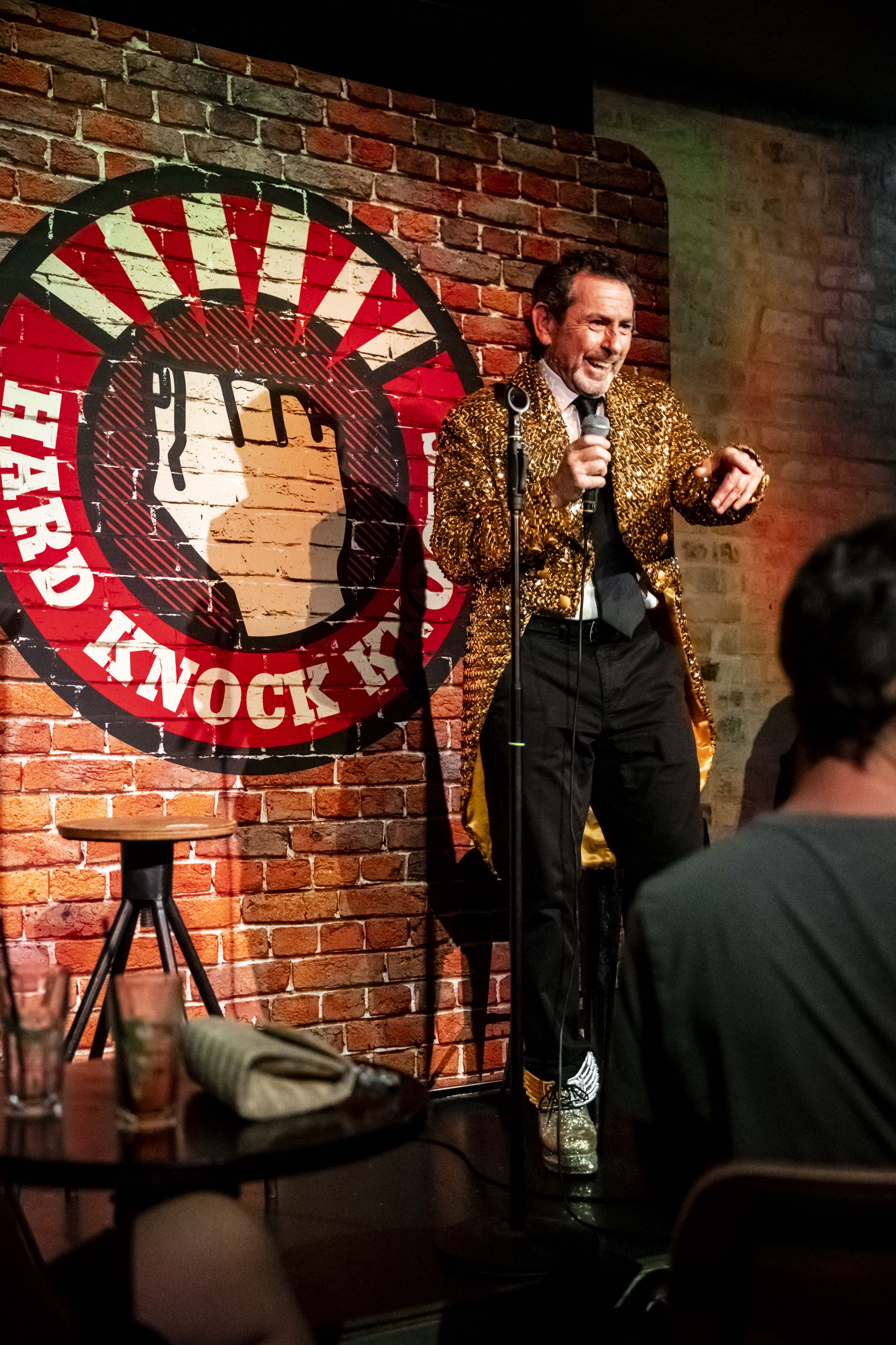 Nick Abrahams performing on stage at a comedy club in his signature gold jacket